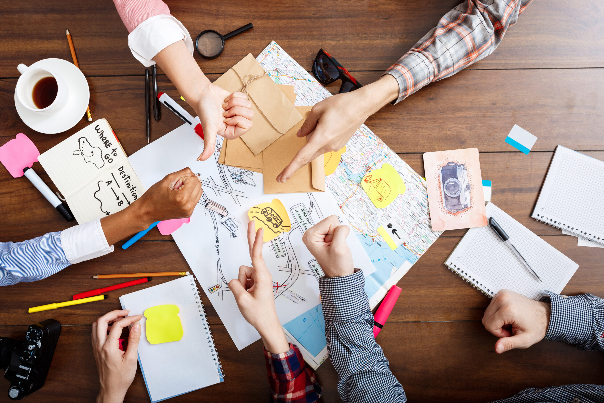 People around a table with innovated decorations, papers, maps, notebooks, pens and post-it-notes. Only hands are visible and they are playing Rock, Paper, Scissors-game.